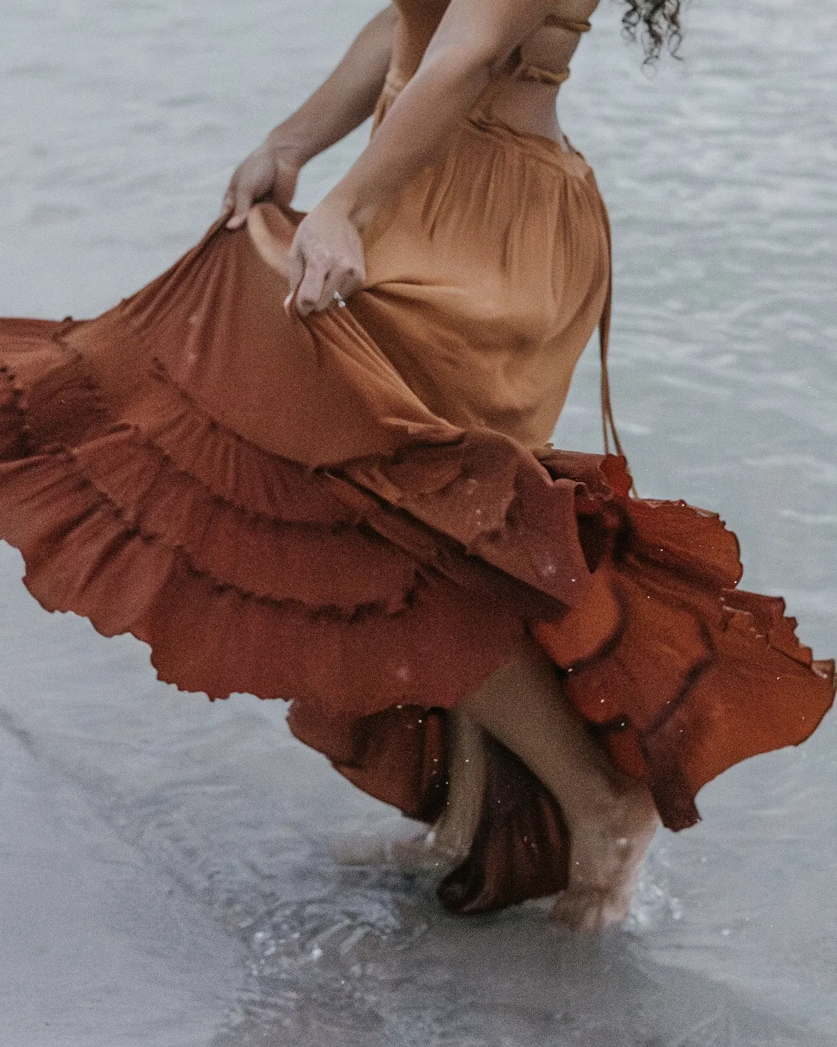 Audra Espinoza, founder of Lu PR, in a flowing terracotta dress walking through soft ocean water at golden hour