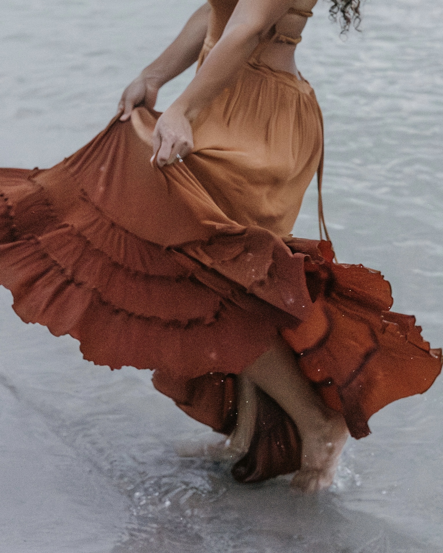 Editorial faceless portrait of a woman in a flowing rust dress walking through water